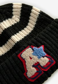 Gestreifte schwarze und cremefarbene Strickmütze mit einem großen bestickten Patch, rotem Hintergrund mit einem strukturierten cremefarbenen Buchstaben "A" und blauen Sternen.