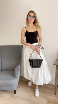Top preto sem mangas combinado com uma saia maxi branca e fluida. Segura uma mala preta texturizada e usa óculos de sol redondos com lentes azuis.