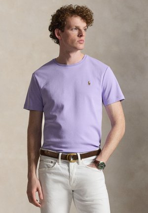 Mann mit lockigem Haar, der ein fliederfarbenes kurzärmeliges T-Shirt, weiße Hose, braunen Gürtel und Armbanduhr trägt, schaut zur Seite.