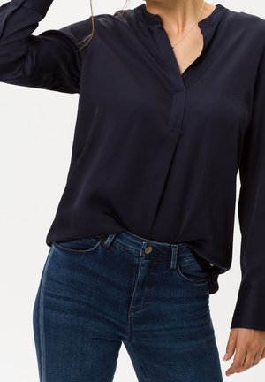 Woman wearing a black long-sleeve V-neck blouse tucked into dark blue denim jeans with a silver necklace visible.