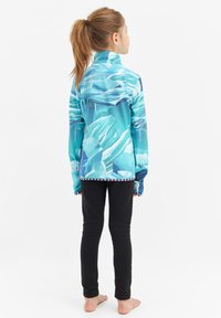 Färgglad, långärmad jacka i nyanser av blått och turkos med ett abstrakt mönster. Högt halsbande, kombinerad med svarta tights.