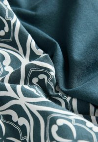 Textil mit einem türkisfarbenen Hintergrund und einem weißen, filigranen Blumenmuster. Weiche Textur, glattes Gewebe mit hängenden Falten.