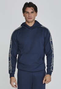 Felpa blu navy con tasca a marsupio e cappuccio regolabile. Presenta accenti del logo beige lungo le maniche. Realizzata in tessuto morbido e elasticizzato.