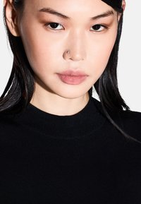 Top in maglia nera con scollo alto. Primo piano di un volto con pelle liscia, trucco sottile e un piercing al narice. Capelli scuri, texture liscia.