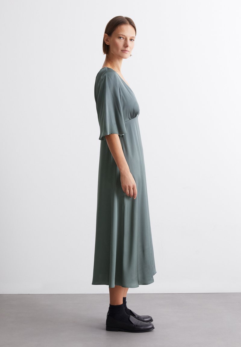 Grünes Midi-Kleid aus glattem, leichtem Stoff mit tiefem V-Ausschnitt und lockeren Ärmeln. Schwarze flache Schuhe runden den Look ab.