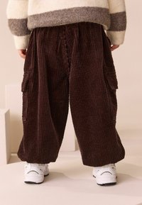 Bruna kordflrossover med vida ben, med sidofickor och en strukturerad tyg. Kombineras med vita sneakers för en avslappnad look.