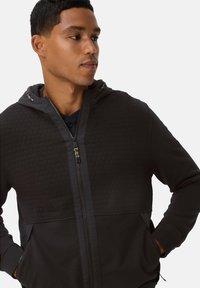 Sweat à capuche zippé noir avec une texture en nid d'abeille sur la partie supérieure, section inférieure unie, capuche ajustable et poches avant. Tissu lavable en machine.