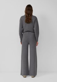 Woman with long wavy brown hair wearing a loose grey sweater and wide-legged grey pants, standing against a plain light grey background.