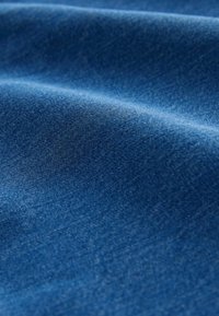 Gładka, niebieska tkanina dżinsowa z widoczną diagonalną strukturą splotu oraz delikatnymi fałdami tworzącymi miękkie cienie.