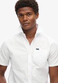 Camisa blanca de manga corta con botones, bolsillo frontal y cuello abotonado. La tela parece suave con un ligero brillo. Etiqueta de la marca en azul en el bolsillo.