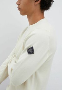 Maglione lavorato a maglia color crema con colletto rotondo e un'etichetta logo triangolare sulla manica sinistra. Texture morbida con vestibilità rilassata.