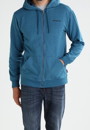 Mężczyzna w niebieskiej bluzie Patagonia z zamkiem, mający ręce w przednich kieszeniach, ubrany w ciemnoniebieskie dżinsy na jasnym, jednolitym tle.