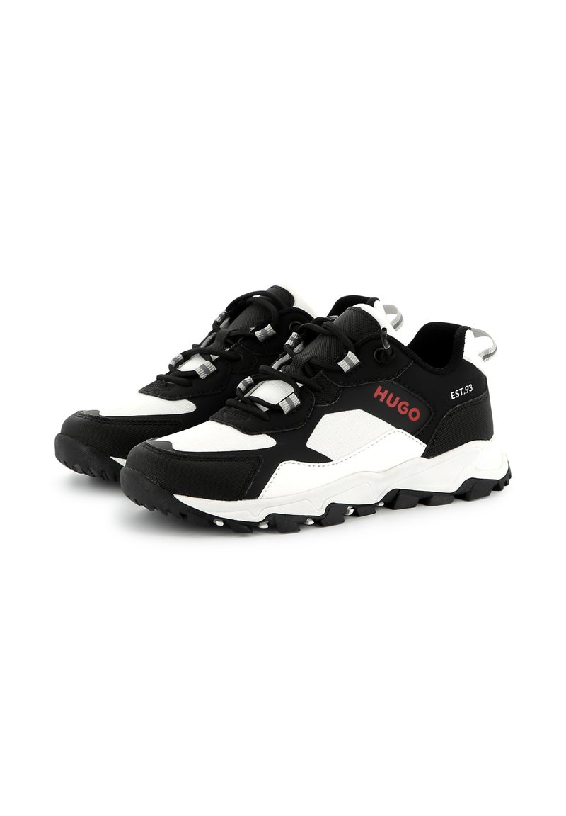 Scarpe sportive in bianco e nero con tomaia testurizzata, suola in gomma sagomata e hardware riflettente. Logo sul lato in rosso.