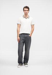 Junger Mann steht mit einer Hand in der Tasche, trägt ein weißes Poloshirt, dunkelgraue Jeans und graue Sneakers vor einem einfarbigen, hellen Hintergrund.