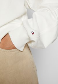Personne portant une chemise blanche à manches longues avec un petit logo rouge et bleu sur le poignet, main dans la poche d’un pantalon beige.