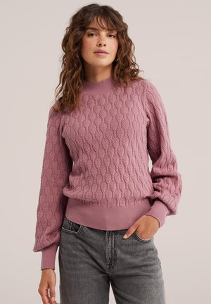 Frau mit lockigem Haar, die einen strukturierten mauvefarbenen Pullover und graue Jeans trägt, posiert mit einer Hand in der Tasche vor einem schlichten Hintergrund.