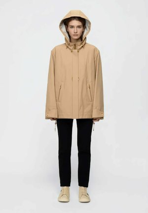 Giovane che indossa una giacca beige con cappuccio, pantaloni neri e sneaker beige, in piedi contro uno sfondo chiaro uniforme.