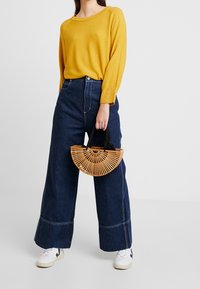 Żółty sweter z prążkowanego dzianiny, ciemne szerokie spodnie dżinsowe z wysokim stanem oraz bambusowa torebka w kształcie wachlarza. Sneakersy z czarnymi akcentami.