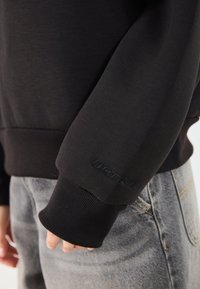 Svart hettegenser med ribbestrikkede mansjetter. Har subtilt hevet merkevarelogo på ermet. Kombinert med lysegrå denimjeans.