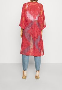 Punainen pitsikimono, jossa on kukkakuvio, läpikuultava kangas, leveät hihat ja nilkkapituus, puettuna sinisten farkkujen ja korkokenkien päälle.