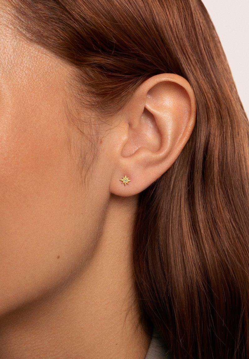 Boucle d'oreille en forme d'étoile dorée avec une surface texturée et une pierre centrale transparente, portée sur l'oreille d'un modèle, avec des cheveux bruns partiellement visibles.