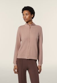 Felpa con zip di colore rosa chiaro, colletto alto, texture morbida, maniche lunghe e taglio corto, abbinata a leggings marroni.