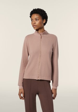 Felpa con zip di colore rosa chiaro, colletto alto, texture morbida, maniche lunghe e taglio corto, abbinata a leggings marroni.