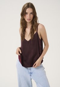 Camisola in satin bordeaux con scollatura a V, spalline sottili e vestibilità abbondante. Abbinata a jeans in denim blu chiaro con leggeri strappi.
