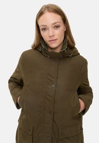 Olijfgroene, geïsoleerde jas met een capuchon, voorzien van knoopsluiting, dubbele voorzakken en een geruite voering. Glad textuur.