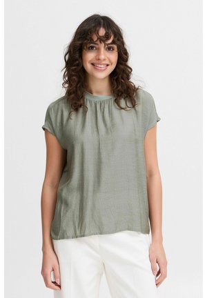 Blusa corta olive green con maniche a raglan e scollatura rotonda, decorata con un dettaglio arricciato. Tessuto leggero e comodo con una leggera texture.