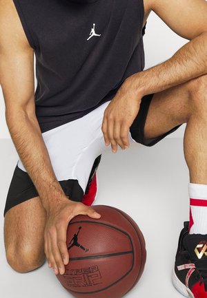 Mann kniet mit einer Hand auf einem Basketball, trägt ein schwarzes ärmelloses Shirt, weiße und schwarze Shorts, weiße Socken und schwarz-rote Sneakers.