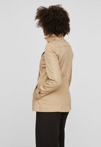 Beige waterdichte jas met een hoge kraag, aansluitend ontwerp, zijzakken en knopenaccenten. Gladde textuur, halflang.