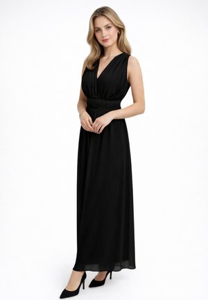 Kobieta o blond włosach, ubrana w czarną, długą suknię bez rękawów i czarne szpilki, pozująca na białym tle.