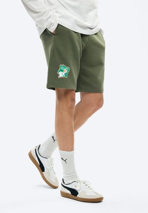 Άνδρας που φοράει χακί πράσινες σορτς με πατς της Ακτής Ελεφαντοστού, λευκές κάλτσες Puma και λευκά αθλητικά παπούτσια Puma με μαύρες λεπτομέρειες, στέκεται με τα χέρια στις τσέπες.