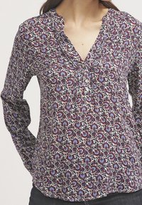 Blouse à manches longues avec un motif floral aux teintes de bordeaux, bleu et crème. Col en V avec des plis et des boutons décoratifs à l'avant.