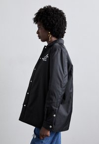 Zwarte waterdichte jas met drukknoopsluiting, opstaande kraag, logo op de borst en een losse pasvorm. Gekleed met blauwe jeans en een opvallende oorbel.