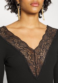 Top preto com um decote em V profundo, apresentando detalhes intrincados em renda preta ao longo da gola, com uma textura de tecido em nervuras contrastantes.