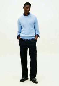 Šviesiai mėlynas džemperis uždėtas ant dryžuoto mėlynai baltos spalvos marškinio, derinamas su tamsiomis kelnėmis ir juodais batais. Paprastas, klasikinis stilius ir pritaikymas.