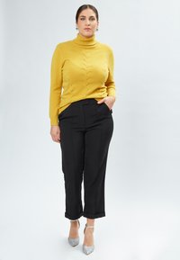 Suéter de cuello vuelto amarillo con patrón de punto, combinado con pantalones negros recortados. La modelo lleva tacones plateados brillantes. Fondo limpio.