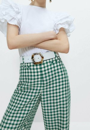 Person med armene krydset iført hvidt kortærmet top med flæser, grønne og hvide ternede bukser og hvidt bælte med stor sekskantet spænde.