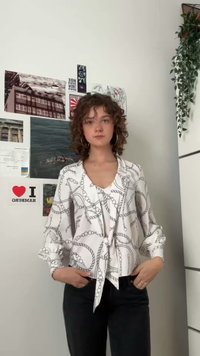 Hvid bluse med kædemønster, lange ærmer og bundet foran. Kombineret med mørke jeans. Krøllet hår og hoopsøreringe synlige.