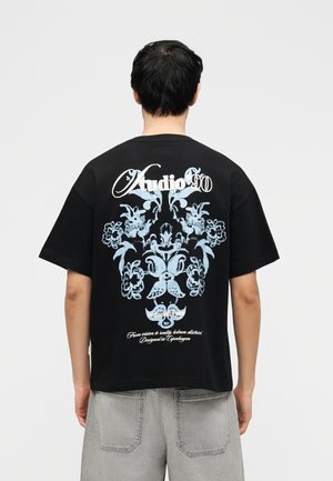 Person trägt ein schwarzes T-Shirt mit blauem Blumenmuster und weißem Schriftzug "Audio 90" und steht vor einem schlichten hellen Hintergrund.