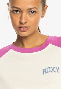 Koszulka RXY w kremowym kolorze z różowymi rękawami raglanowymi; zawiera niebieski tekst logo po lewej stronie; wykonana z miękkiego materiału bawełnianego.