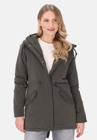 Femeie îmbrăcată într-o geacă parka cu glugă de culoare verde închis, cu buzunare în față, peste un top alb și blugi gri deschis, zâmbind cu părul blond lăsat liber.