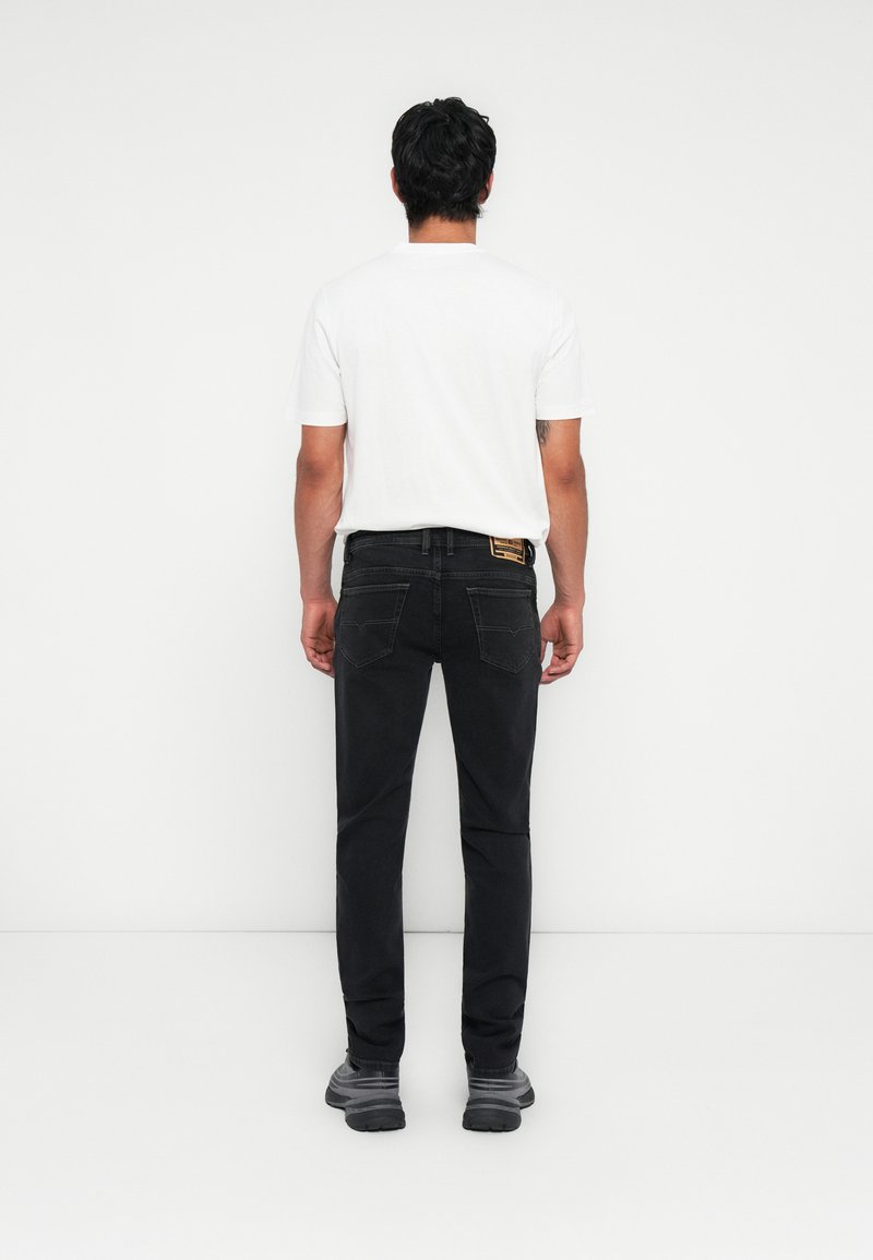Mann mit dem Rücken zur Kamera, trägt ein weißes T-Shirt, schwarze Jeans und graue Turnschuhe, steht vor einem schlichten weißen Hintergrund.