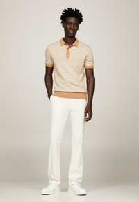 Polo beige a righe con colletto beige, abbinato a pantaloni bianchi e sneakers bianche. Maniche corte e vestibilità rilassata.