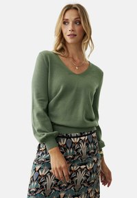 Suéter verde de punto con cuello en V y mangas abullonadas, combinado con una falda de estampado que presenta diseños florales en tonos oscuros.