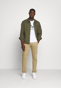 Camisa verde olivo de botones sobre una camiseta blanca con logo, combinada con pantalones beige y zapatillas blancas, de pie sobre un fondo gris claro.