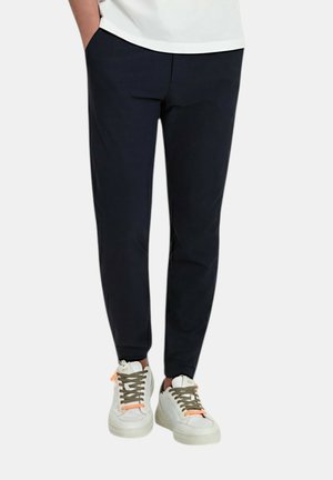 Persona che indossa pantaloni neri affusolati e sneakers bianche con lacci marroni, in piedi con le mani in tasca davanti a uno sfondo semplice.
