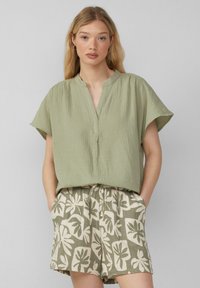 Top corto color verde oliva con maniche corte, scollo a V e tessuto strutturato, abbinato a pantaloni beige con un motivo di foglie verde.